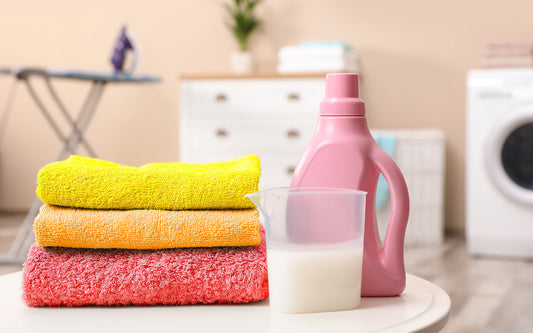 folded yellow, orange, and pink towels and a small amount of a detergent with the detergent bottle on the table while an ironing table, a dresser and a washing machine are appearing behind them